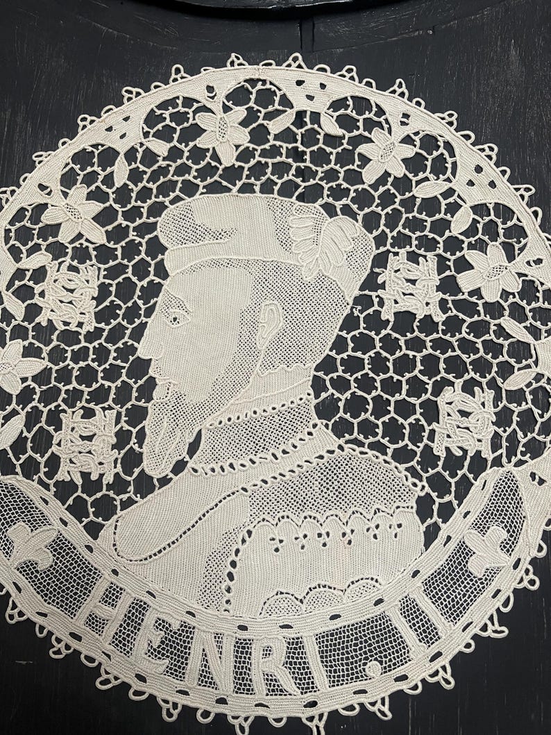 May include: Cream-colored, circular lace doily featuring a portrait of a man, possibly historical, with the name "HENRI" around the border. The lace has a delicate, openwork design with floral and geometric patterns.