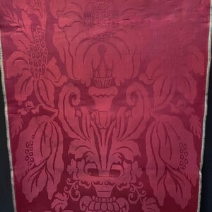 May include: A close-up of a deep red fabric with a damask pattern of stylized flowers and leaves. The fabric has a subtle sheen and a textured surface.