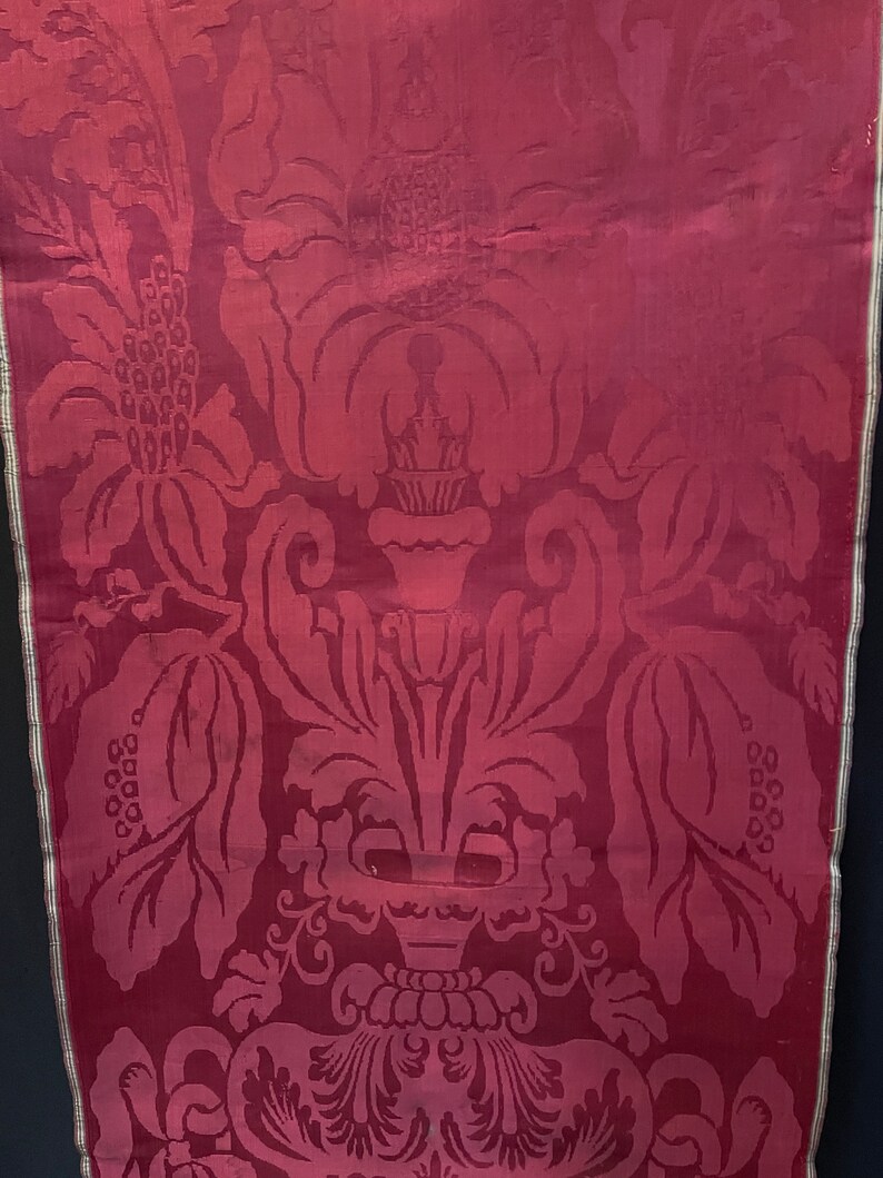 May include: A close-up of a deep red fabric with a damask pattern of stylized flowers and leaves. The fabric has a subtle sheen and a textured surface.