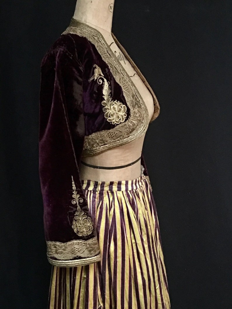 May include: A burgundy velvet cropped jacket with gold embroidery and a matching striped skirt. The jacket has a V-neckline and long sleeves. The skirt is a full skirt with a high waist.
