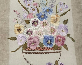 May include: Embroidered floral artwork featuring a variety of colorful flowers in a vase. The flowers include pansies, daisies, and other blooms in shades of purple, blue, pink, and yellow. The vase is a light beige color with a brown checkered pattern.