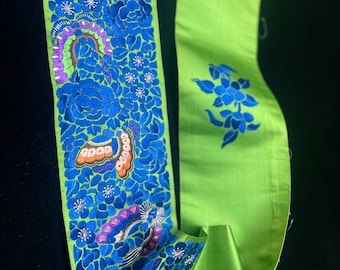 May include: A vibrant green satin ribbon with intricate embroidery. The ribbon features a detailed design of blue flowers and butterflies with orange and purple accents. The design is set against a bright green background, creating a striking contrast.