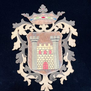 Op de afbeelding: Een vintage geborduurd wapenschild met een bruin kasteel met een rode deur en gele bakstenen. Het wapenschild is omgeven door een sierlijk gouden en zilveren frame met een kroon bovenaan.