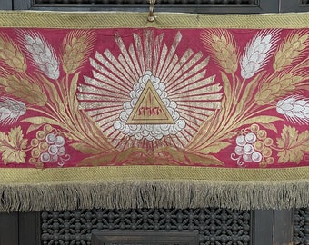 May include: A red fabric banner with gold and silver embroidery. The banner features a triangle with the Hebrew word "שלום" (peace) inside. The banner also has wheat stalks and grapes on either side of the triangle.
