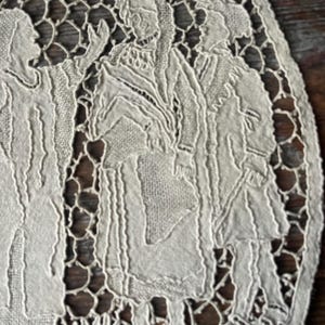 May include: Close-up of a cream-colored, oval-shaped lace doily. The doily features a detailed design of figures in period clothing, with a delicate openwork border. The background is a dark wood surface.
