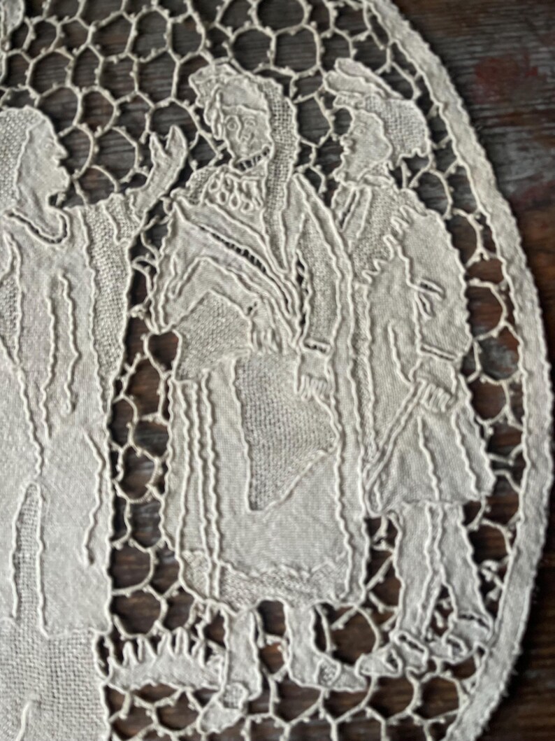 May include: Close-up of a cream-colored, oval-shaped lace doily. The doily features a detailed design of figures in period clothing, with a delicate openwork border. The background is a dark wood surface.