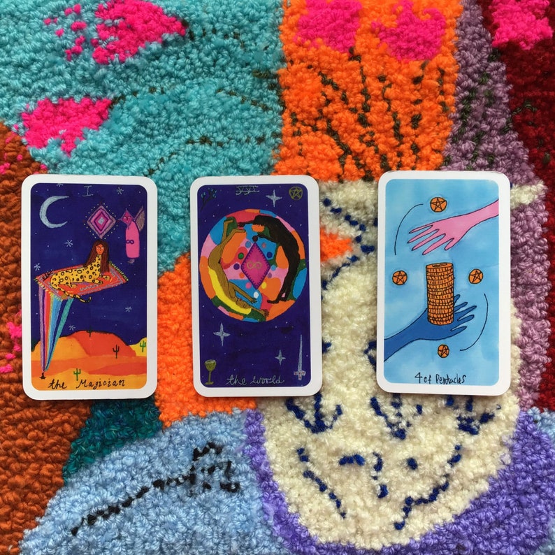 Puede incluir: Tres cartas de tarot con ilustraciones coloridas se muestran sobre una alfombra texturizada y multicolor. Las cartas presentan im&aacute;genes de "El Mago", "El Mundo" y "4 de Oros". La obra de arte incluye elementos celestiales, figuras y formas geom&eacute;tricas, con texto en la parte inferior de cada carta.