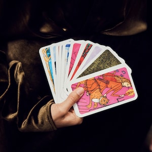 Puede incluir: Una mano sosteniendo un abanico de cartas del tarot. Cada carta presenta ilustraciones coloridas con un borde blanco. Las cartas tienen una combinaci&oacute;n de colores rosa, naranja, azul y verde. El fondo es oscuro.