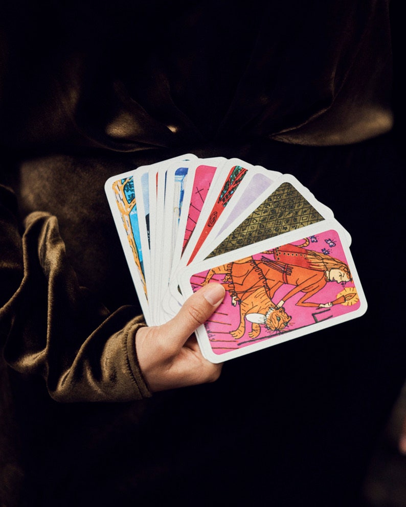Puede incluir: Una mano sosteniendo un abanico de cartas del tarot. Cada carta presenta ilustraciones coloridas con un borde blanco. Las cartas tienen una combinaci&oacute;n de colores rosa, naranja, azul y verde. El fondo es oscuro.