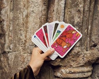 May include: A hand holding a deck of tarot cards with colorful illustrations. The cards feature images of people, animals, and celestial bodies, set against a pink background. The cards are fanned out, revealing their unique designs.