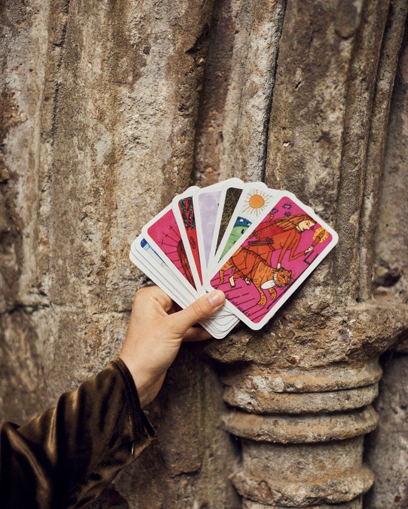 Puede incluir: Una mano sosteniendo una baraja de cartas del tarot con ilustraciones coloridas. Las cartas presentan im&aacute;genes de personas, animales y cuerpos celestes, sobre un fondo rosa. Las cartas est&aacute;n abanicadas, revelando sus dise&ntilde;os &uacute;nicos.