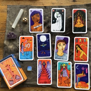 Puede incluir: Una baraja de cartas del tarot con ilustraciones coloridas, que incluyen un unicornio, una persona meditando y una figura con alas. La baraja se muestra con cristales y una caja sobre una superficie de madera.