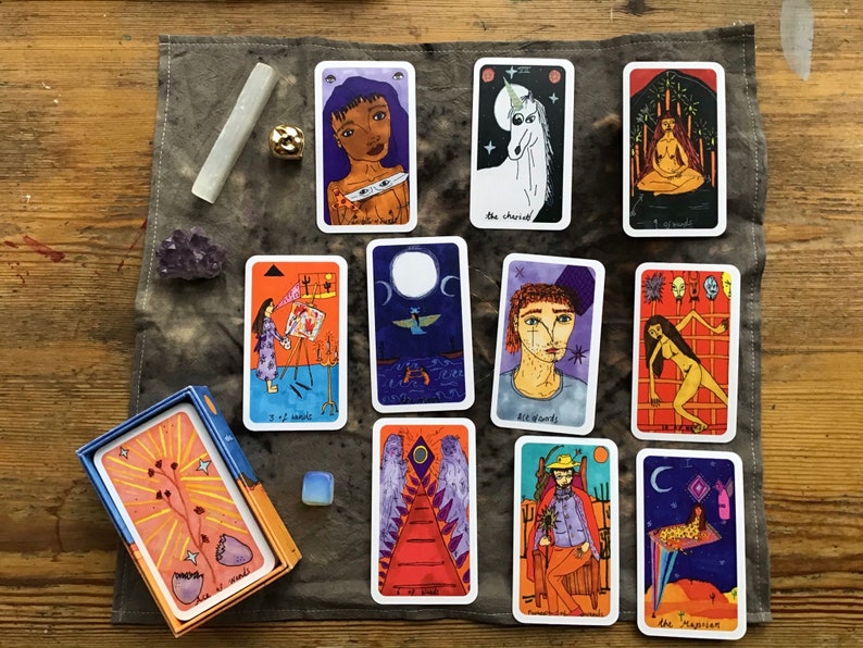 Puede incluir: Una baraja de cartas del tarot con ilustraciones coloridas, que incluyen un unicornio, una persona meditando y una figura con alas. La baraja se muestra con cristales y una caja sobre una superficie de madera.