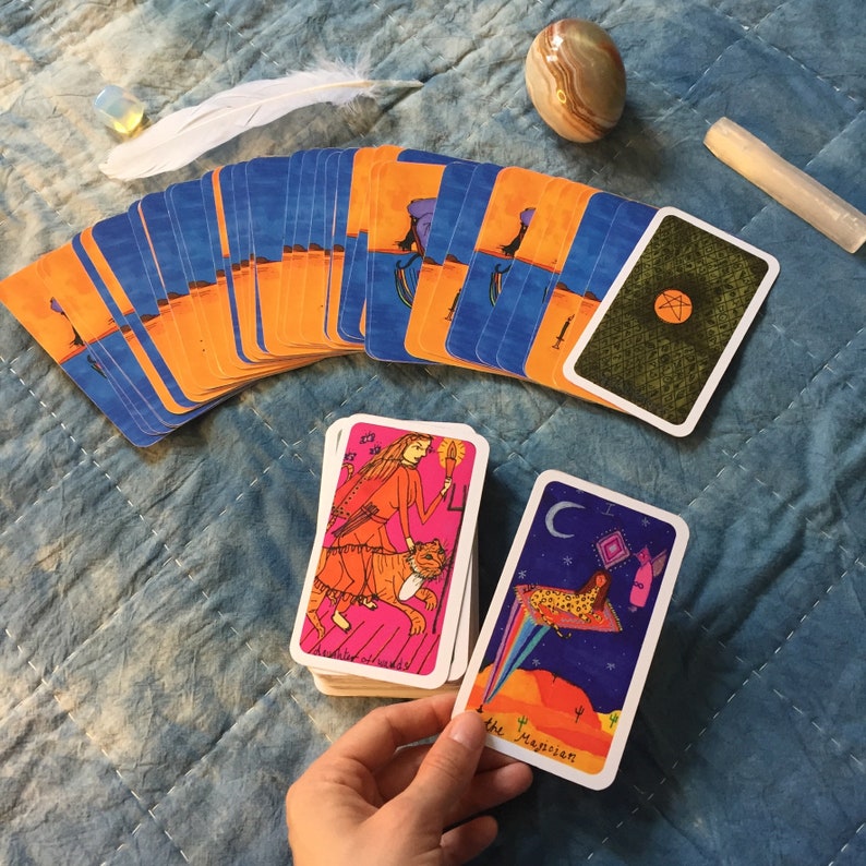Puede incluir: Una baraja de cartas del tarot con dorsos naranjas y azules abanicados sobre una superficie acolchada azul. Dos montones de cartas est&aacute;n en primer plano, con una carta sostenida por una mano. Las cartas presentan ilustraciones coloridas. Tambi&eacute;n hay una pluma, cristales y una piedra.