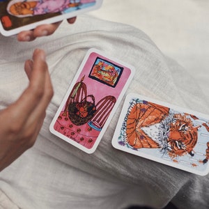 Puede incluir: Cartas de tarot con ilustraciones de un tigre y una escena interior rosa. Las cartas son rectangulares con bordes blancos. Una carta est&aacute; sostenida en la mano de una persona. La otra carta est&aacute; sobre una superficie blanca.