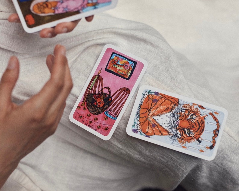 Puede incluir: Cartas de tarot con ilustraciones de un tigre y una escena interior rosa. Las cartas son rectangulares con bordes blancos. Una carta est&aacute; sostenida en la mano de una persona. La otra carta est&aacute; sobre una superficie blanca.