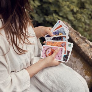 Puede incluir: Una persona sostiene una baraja de cartas de tarot coloridas. Cada carta presenta ilustraciones &uacute;nicas con varios colores, incluyendo rosa, azul y naranja. Las cartas son rectangulares y tienen esquinas redondeadas. El fondo est&aacute; borroso, con follaje verde y un muro de piedra.