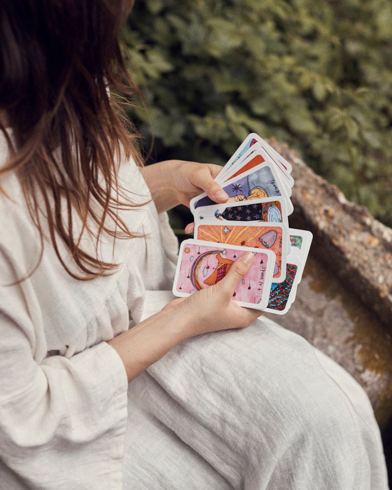 Puede incluir: Una persona sostiene una baraja de cartas de tarot coloridas. Cada carta presenta ilustraciones &uacute;nicas con varios colores, incluyendo rosa, azul y naranja. Las cartas son rectangulares y tienen esquinas redondeadas. El fondo est&aacute; borroso, con follaje verde y un muro de piedra.