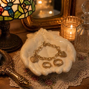 May include: A white ceramic dish shaped like two cupped hands holding a gold-toned chain link bracelet with two heart-shaped charms and two matching gold-toned bands. The jewelry rests centrally within the palm-shaped vessel, displaying a metallic finish.