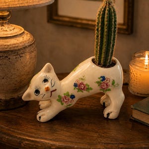 May include: A white ceramic cat-shaped planter with a cactus inside. The planter is decorated with pink and blue floral patterns. The cat is in a playful pose, with its front paws extended. The planter is on a wooden surface.