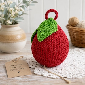 May include: A red crochet string holder shaped like a fruit with a green leaf and a small red ring at the top. A tan tag with the text Handmade Vintage String Holder and a small heart icon is placed next to the item on a white lace doily.