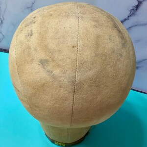May include: A beige canvas-covered mannequin head with visible seams running vertically across the surface. The fabric shows signs of wear with scattered dark spots and discolouration. A small yellow label at the base displays the text size 22.