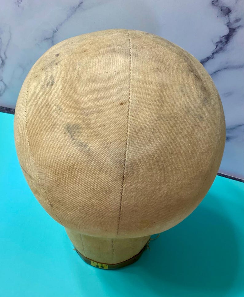 May include: A beige canvas-covered mannequin head with visible seams running vertically across the surface. The fabric shows signs of wear with scattered dark spots and discolouration. A small yellow label at the base displays the text size 22.