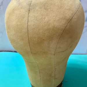 May include: A beige canvas-covered millinery head form with visible vertical seams and a brown base. A small rectangular label on the base clearly displays the text 'SIZE 22'. The surface of the fabric shows light signs of wear and discolouration.