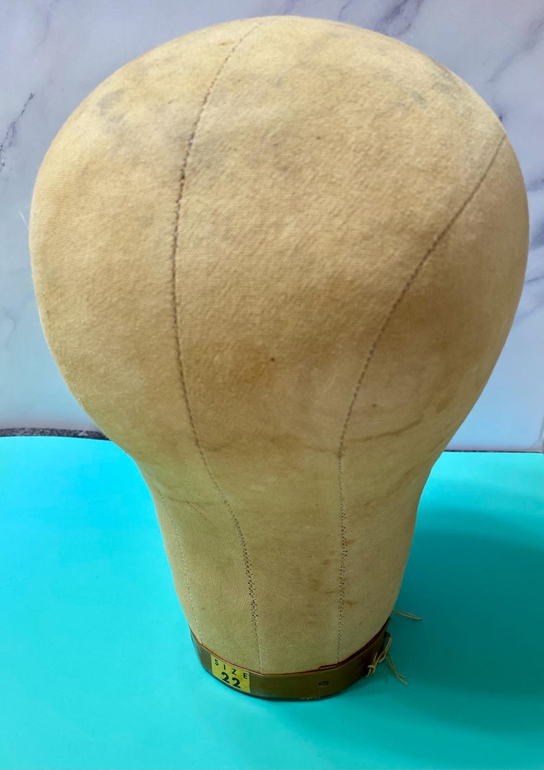 May include: A beige canvas-covered millinery head form with visible vertical seams and a brown base. A small rectangular label on the base clearly displays the text 'SIZE 22'. The surface of the fabric shows light signs of wear and discolouration.