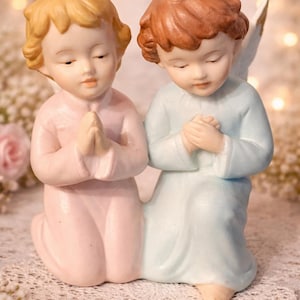 May include: A ceramic figurine of two kneeling angels with clasped hands. One angel wears a pale pink robe, the other a pale blue robe. Both have closed eyes and light brown hair. The background has a soft bokeh effect.
