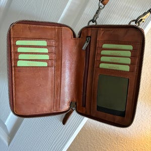 Puede incluir: Una cartera de cuero marr&oacute;n con cierre de cremallera, que presenta m&uacute;ltiples ranuras para tarjetas en ambos lados, una ventana transparente para identificaci&oacute;n y un compartimento vertical con cremallera. Incluye una correa extra&iacute;ble con clips.