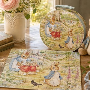 May include: A round tin suitcase and a rectangular jigsaw puzzle featuring Peter Rabbit illustrations. The artwork depicts rabbits in a garden setting with soft green and brown tones. A plush rabbit toy sits beside the tin, which has a light blue handle.