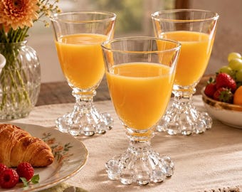 May include: Three clear glass goblets with textured, beaded bases, each filled with bright orange juice. The glasses feature a smooth, tapered bowl and a thick, decorative stem with a cluster of glass spheres at the bottom.