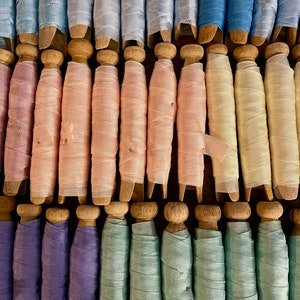 May include: A collection of vintage spools, each with a wooden base and a wrapped ribbon in various pastel colors. The ribbons include shades of blue, pink, green, and purple. The spools are arranged in neat rows, creating a visually appealing display.