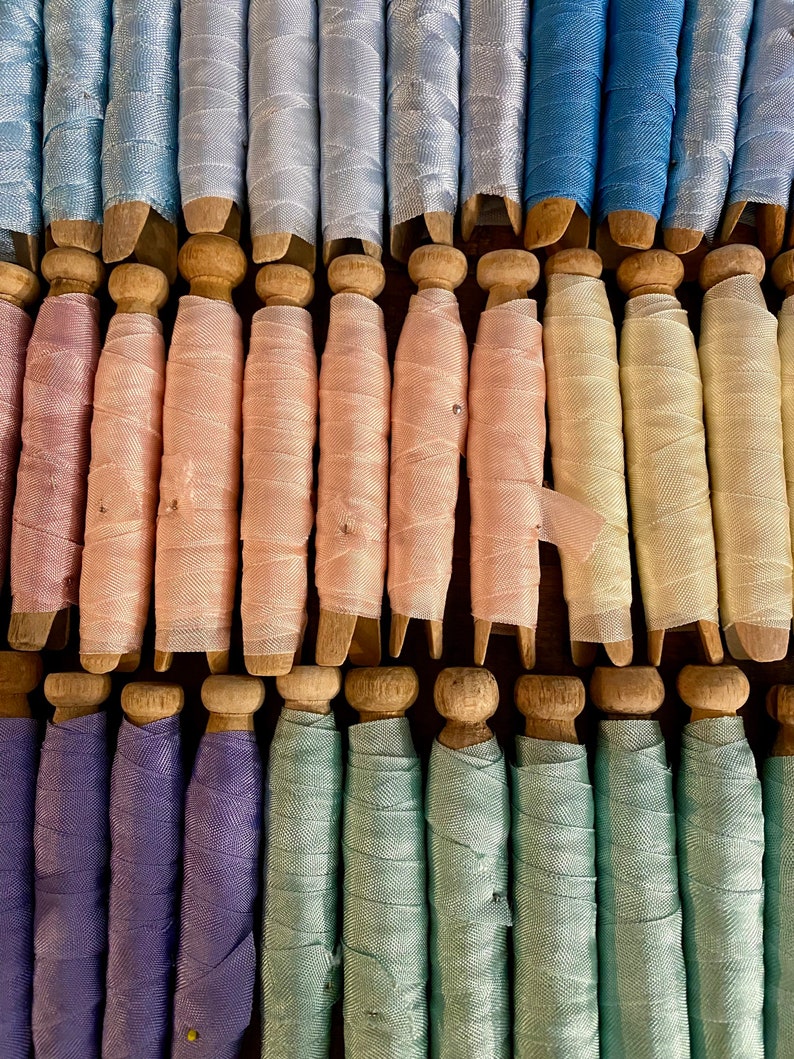 May include: A collection of vintage spools, each with a wooden base and a wrapped ribbon in various pastel colors. The ribbons include shades of blue, pink, green, and purple. The spools are arranged in neat rows, creating a visually appealing display.