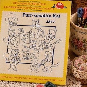 May include: A yellow and white package titled "Purr-sonality Kat" with illustrations of cross-stitch cats. The package includes text in English, French, and Spanish. Embroidery floss, a pincushion, and scissors are also visible.