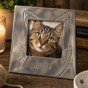 May include: A square silver-toned metal picture frame featuring embossed cat ears at the top corners, etched whiskers on the sides, and a fish skeleton design at the bottom. The frame holds a photograph of a brown tabby cat with green eyes.