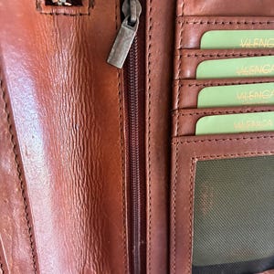 Puede incluir: Una cartera de cuero marr&oacute;n con un compartimento vertical con cremallera y tirador de metal. El interior incluye varias ranuras horizontales para tarjetas con tarjetas de color verde claro etiquetadas VILENCA y un bolsillo de malla.