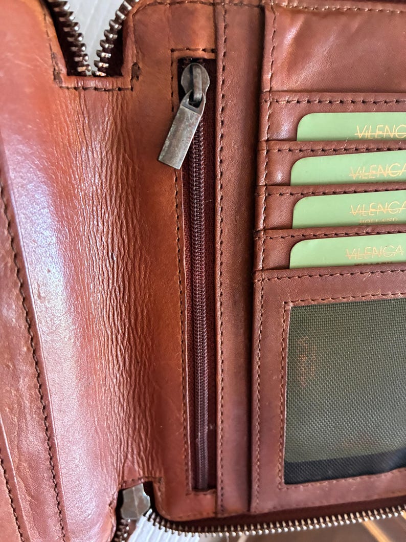 Puede incluir: Una cartera de cuero marr&oacute;n con un compartimento vertical con cremallera y tirador de metal. El interior incluye varias ranuras horizontales para tarjetas con tarjetas de color verde claro etiquetadas VILENCA y un bolsillo de malla.