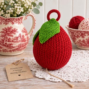 May include: A red crocheted apple-shaped string holder with a green leaf and a small circular loop at the top. A light brown paper tag is attached, featuring the printed text: Handmade Vintage String Holder with a small heart icon below.
