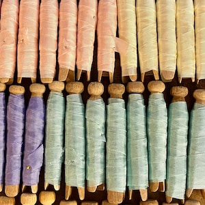 May include: A collection of spools wrapped in various colors of ribbon, including light blue, pink, yellow, green, and burgundy. The spools are made of wood and arranged in neat rows, creating a visually appealing display.