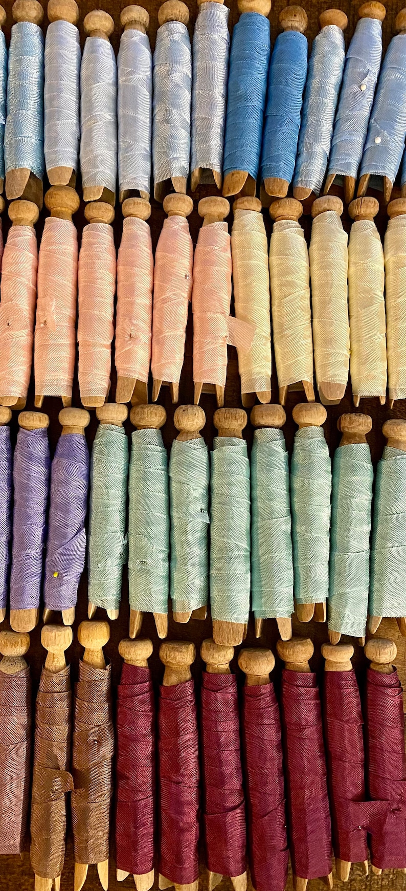May include: A collection of spools wrapped in various colors of ribbon, including light blue, pink, yellow, green, and burgundy. The spools are made of wood and arranged in neat rows, creating a visually appealing display.