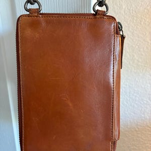 Puede incluir: Un bolso bandolera rectangular de cuero color co&ntilde;ac con cierre de cremallera vertical, acabado liso con sutiles ara&ntilde;azos superficiales y herrajes de metal oscuro, incluyendo dos cierres giratorios superiores unidos a una correa fina.