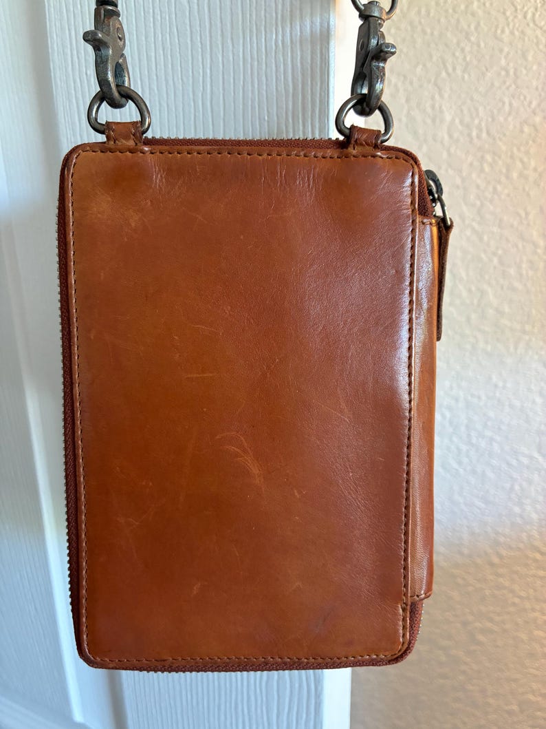 Puede incluir: Un bolso bandolera rectangular de cuero color co&ntilde;ac con cierre de cremallera vertical, acabado liso con sutiles ara&ntilde;azos superficiales y herrajes de metal oscuro, incluyendo dos cierres giratorios superiores unidos a una correa fina.
