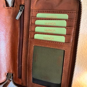 Puede incluir: Una cartera de cuero marr&oacute;n con cierre de cremallera, que presenta m&uacute;ltiples ranuras interiores para tarjetas con tarjetas de color verde claro etiquetadas como Vilenca, un bolsillo vertical con cremallera y una ventana de malla.