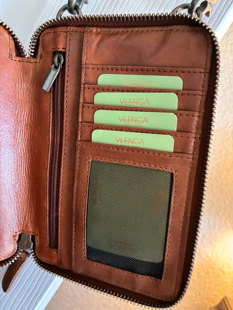 Puede incluir: Una cartera de cuero marr&oacute;n con cierre de cremallera, que presenta m&uacute;ltiples ranuras interiores para tarjetas con tarjetas de color verde claro etiquetadas como Vilenca, un bolsillo vertical con cremallera y una ventana de malla.