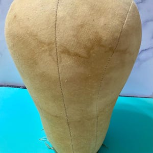 May include: A beige canvas-covered mannequin head with visible vertical seams running from the crown to the base. The head features a smooth, rounded top and a tapered neck area, with a metallic band encircling the bottom edge of the base.
