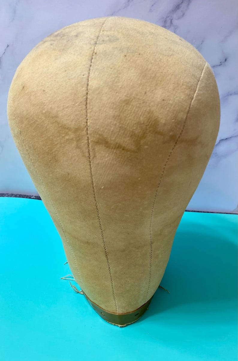 May include: A beige canvas-covered mannequin head with visible vertical seams running from the crown to the base. The head features a smooth, rounded top and a tapered neck area, with a metallic band encircling the bottom edge of the base.