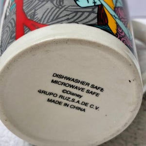Może przedstawiać: Ceramiczny kubek z kolorowym nadrukiem graficznym przedstawiającym postacie z Miasteczka Halloween. Na spodzie kubka znajduje się tekst: DISHWASHER SAFE, MICROWAVE SAFE, Disney, GRUPO RUZ, S.A. DE C.V. oraz MADE IN CHINA.
