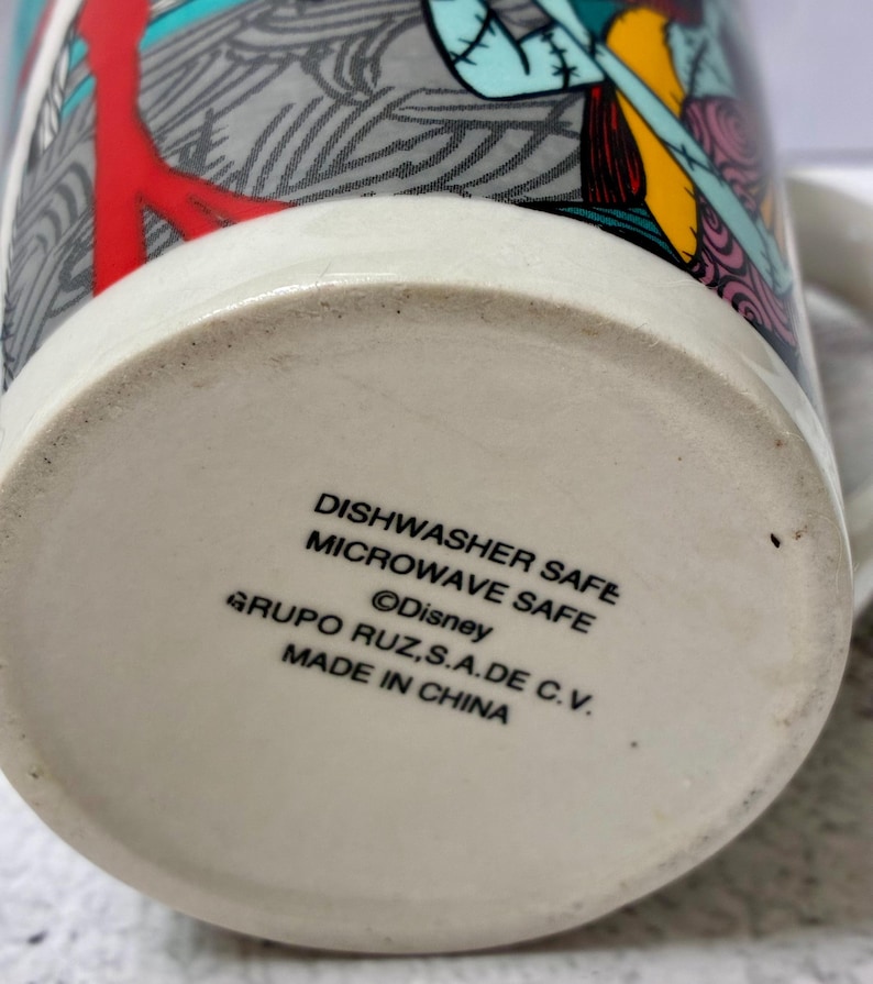 Może przedstawiać: Ceramiczny kubek z kolorowym nadrukiem graficznym przedstawiającym postacie z Miasteczka Halloween. Na spodzie kubka znajduje się tekst: DISHWASHER SAFE, MICROWAVE SAFE, Disney, GRUPO RUZ, S.A. DE C.V. oraz MADE IN CHINA.